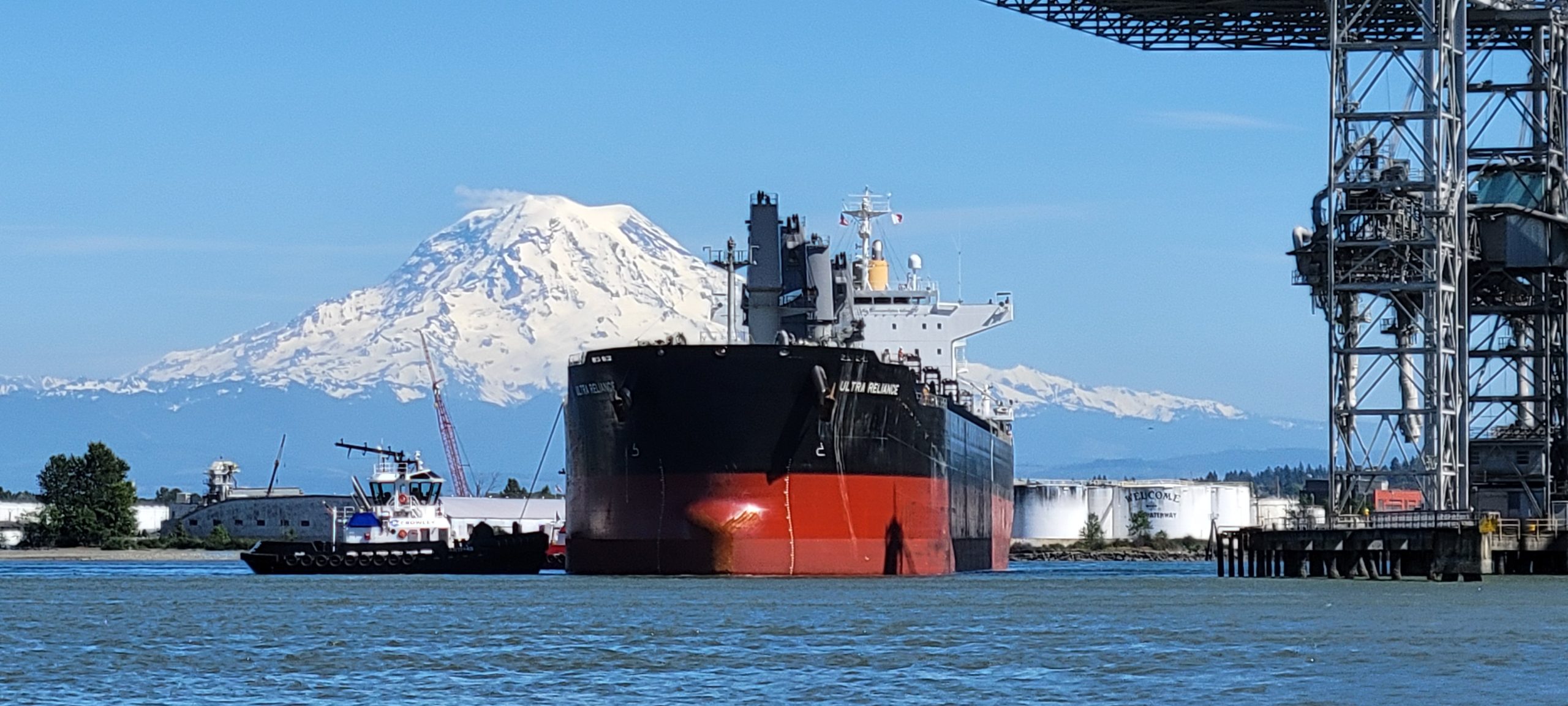 A large cargo ship is docked near an industrial facility with a tugboat beside it and a snow-capped mountain in the background. Plumbers in Black Diamond, WA